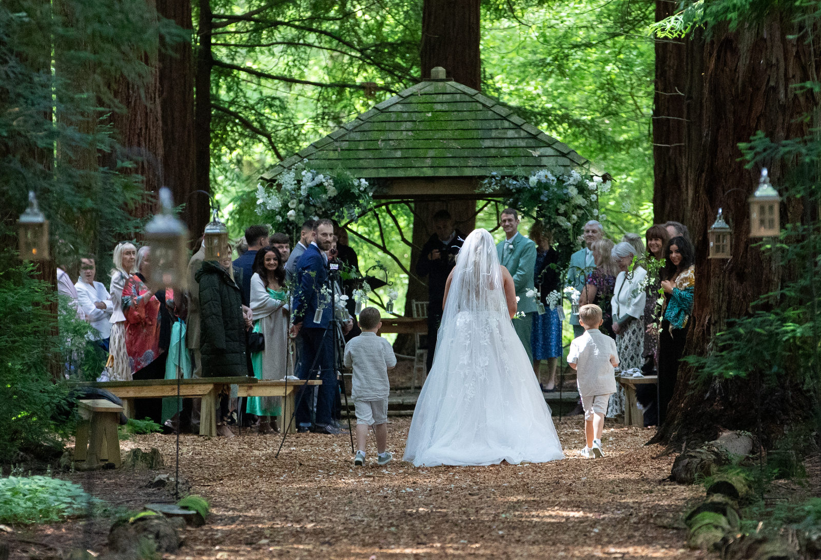 A bride walking through woodland towards an outdoor wedding ceremony