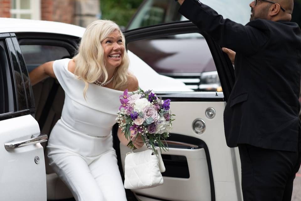 A smiling bride stepping out of a wedding car beneath an umbrella