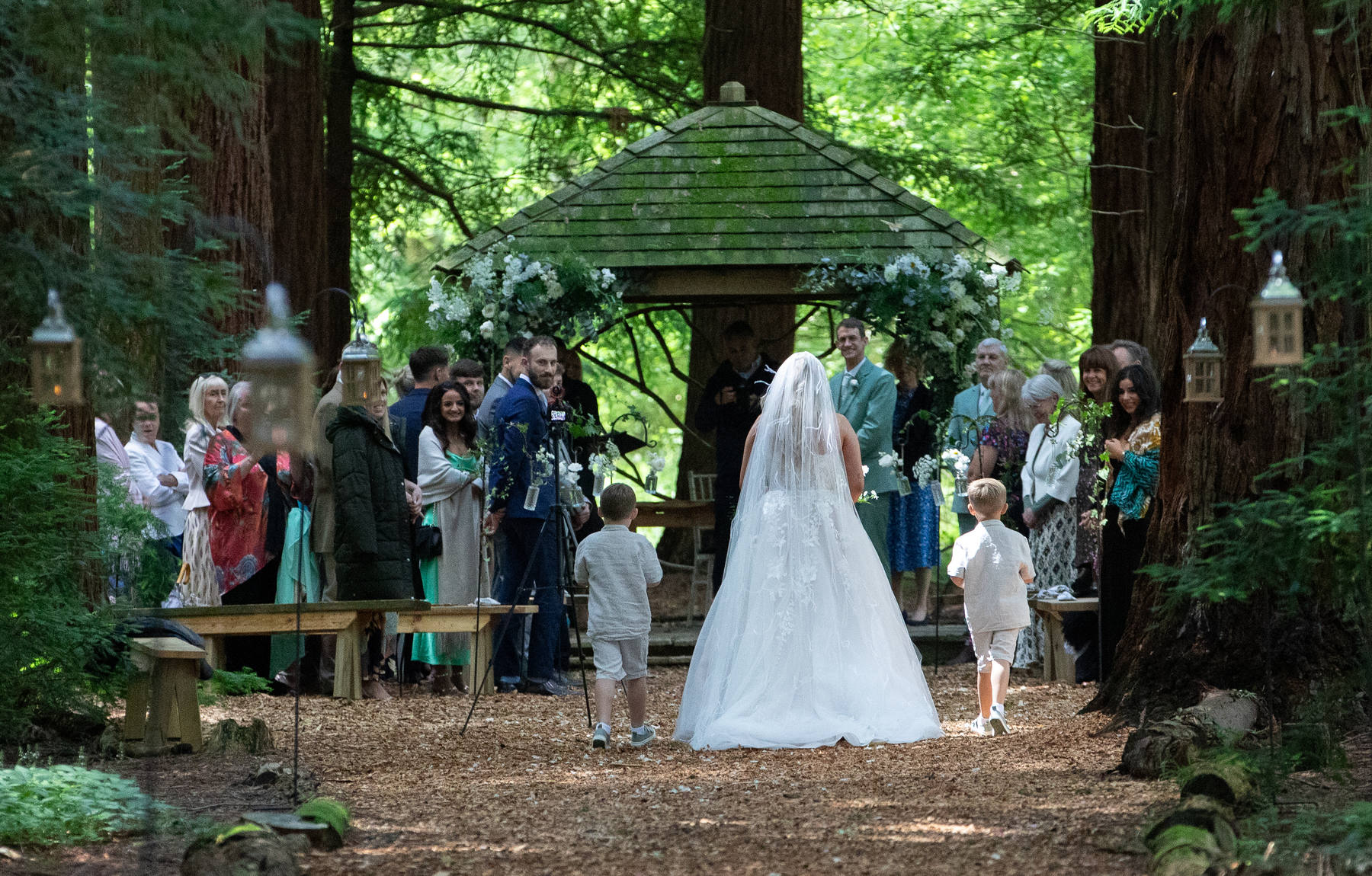 A bride walking through woodland towards an outdoor wedding ceremony
